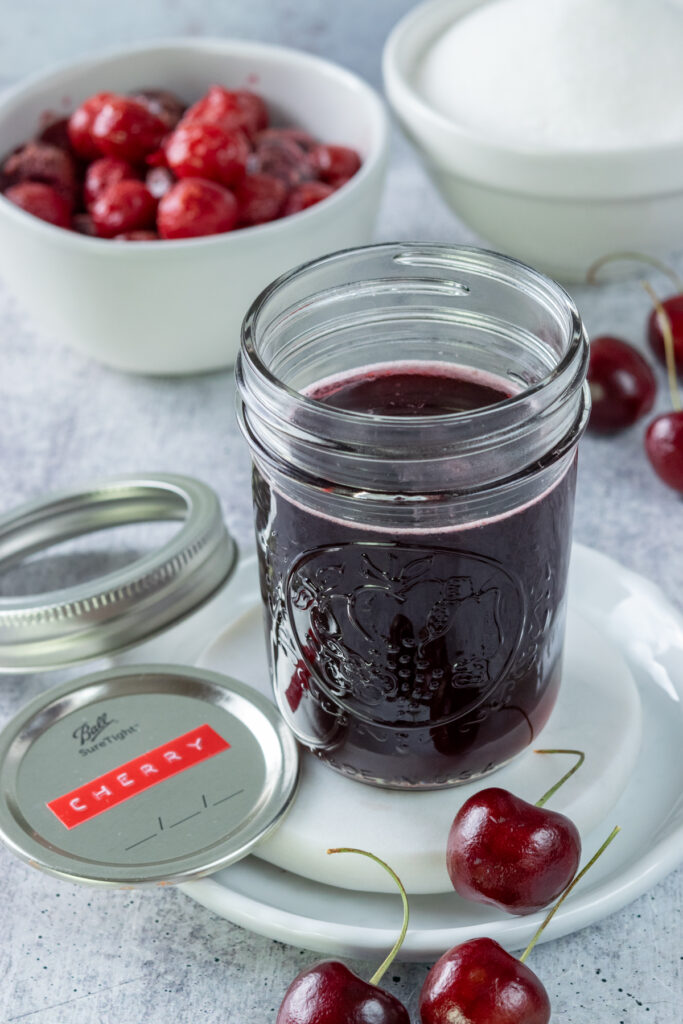 A pint mason jar full of cherry syrup, next to the lid which has a label on it that says, "cherry." The jar of syrup is on a white plate with three fresh cherries on it, and in front of a bowl of frozen cherries and bowl of sugar.