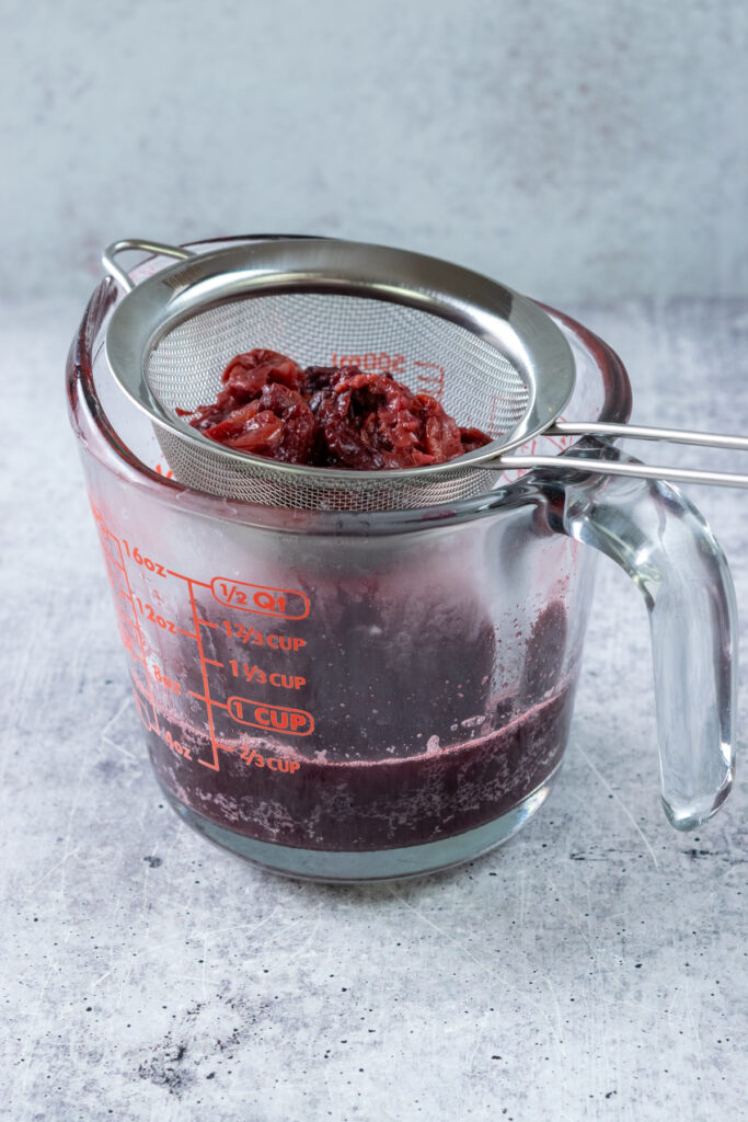 A strainer on top of a glass Pyrex measuring cup. The strainer has the boiled cherries in it and the measuring cup has concentrated cherry juice in it.