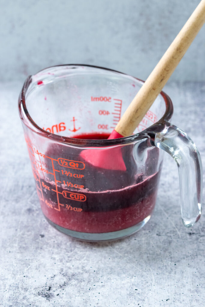A glass measuring cup with cherry juice and sugar added. There's a red rubber spatula in the measuring cup to stir the sugar into the juice.