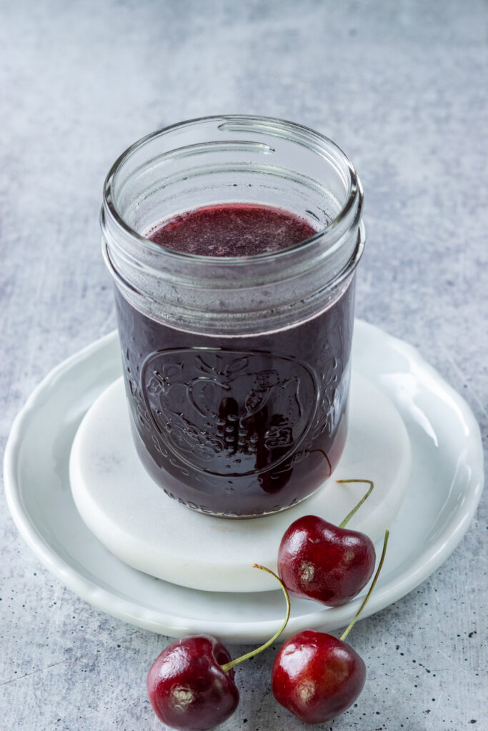 Cherry syrup cooling  in a pint mason jar, with three fresh cherries in front of the jar.