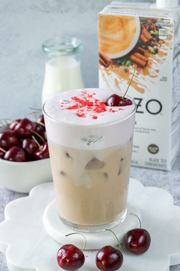 A copycat Starbucks iced cherry chai in a glass showing the layers of the iced chai latte and the cherry cream cold foam on top. The chai drink is in front of a carton of Tazo chai tea concentrate, a small pitcher of milk and a small bowl of cherries.