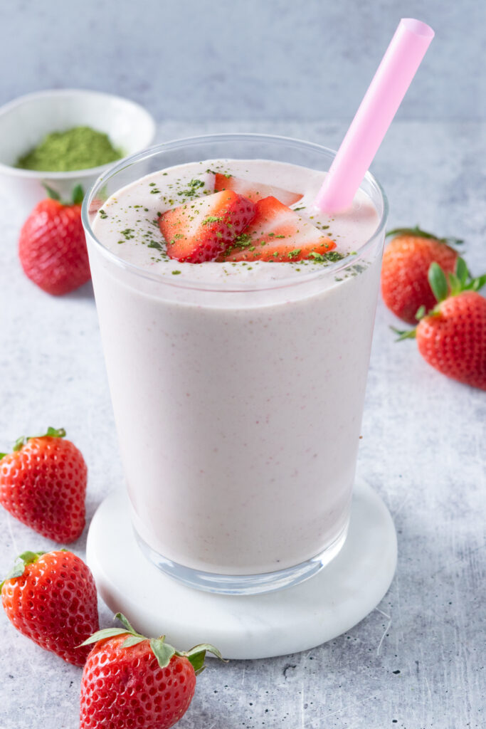 A thick strawberry matcha smoothie in a glass with a pink straw. On top of the smoothie is some sliced strawberries and a sprinkling of matcha powder. There are strawberries scattered around the drink and a small bowl with matcha powder in the background.
