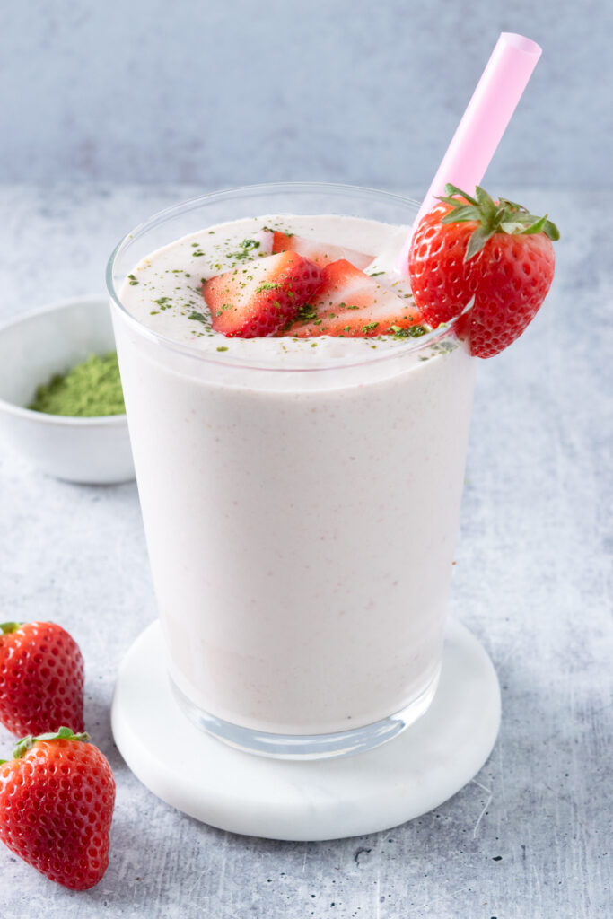 A strawberry matcha smoothie that's in a glass with a pink straw and garnished with a full strawberry on the rim. The smoothie also has a few slices of strawberries on top and is sprinkled with a little bit of matcha powder. Next to the drink, which is on a white coaster, are two strawberries and a small bowl filled with matcha powder.