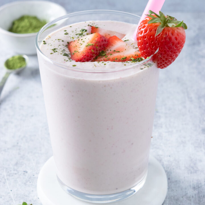A strawberry matcha smoothie with sliced strawberries and matcha sprinkled on top. The smoothie has a pink straw in it and there's a strawberry next to the cup and a small bowl of matcha powder in the background.