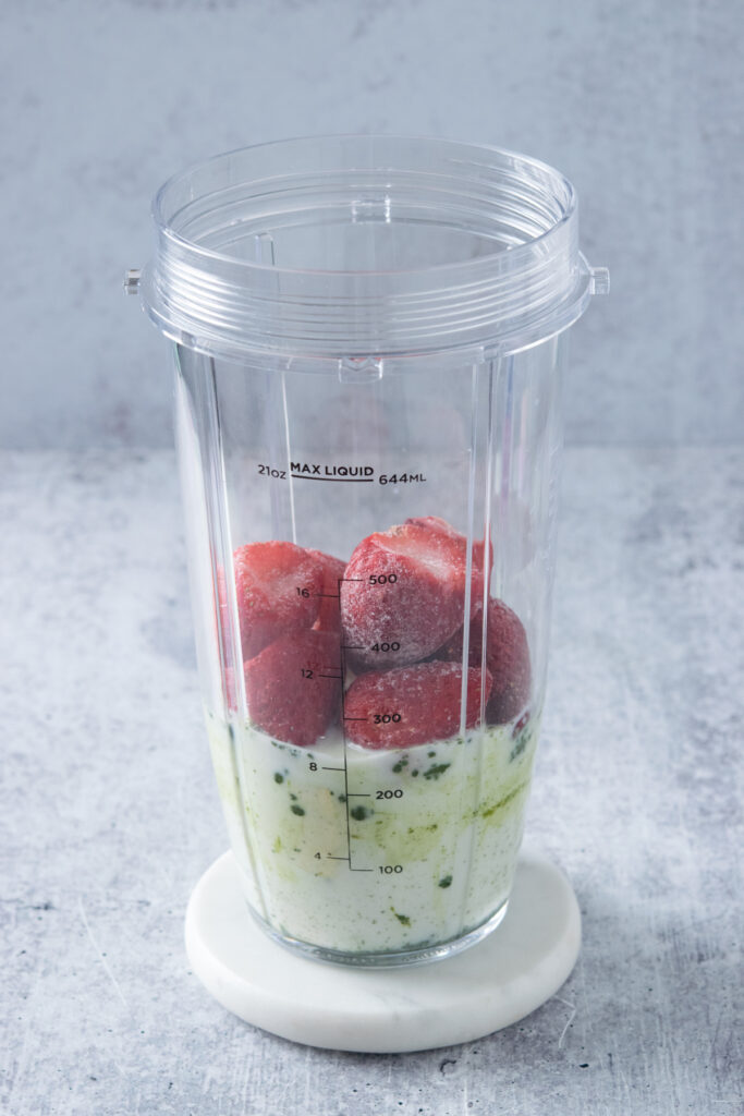 A blender cup with milk, matcha powder, banana and frozen strawberries in it.