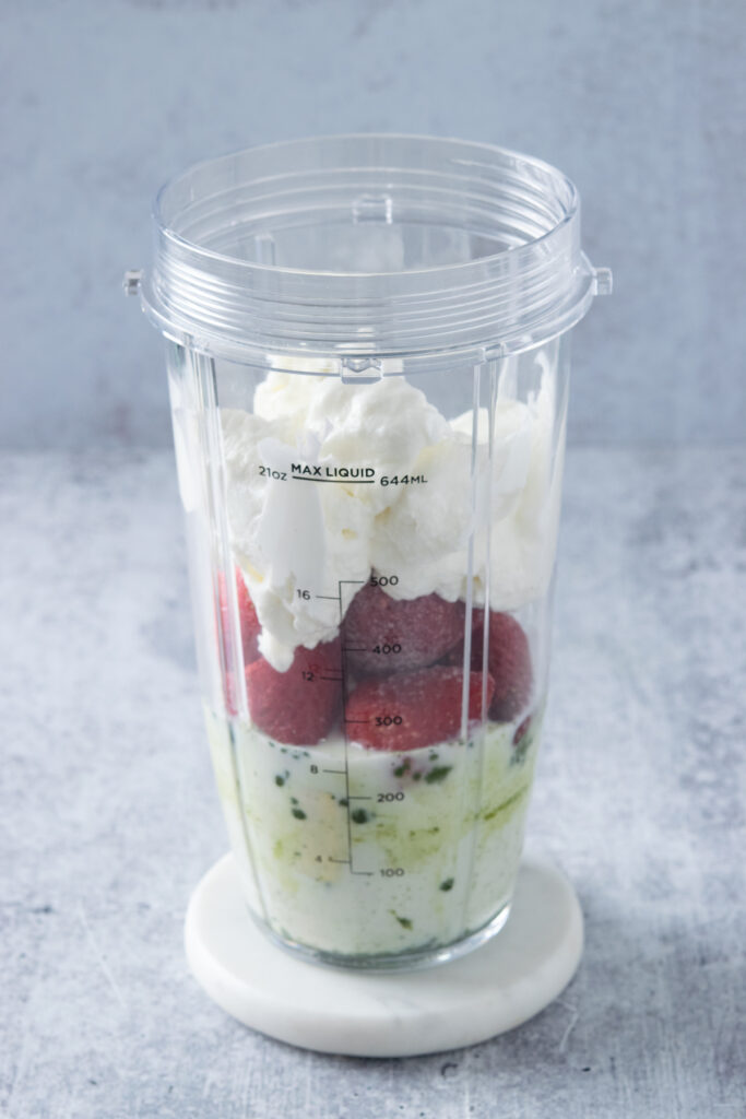 A blender cup with milk, matcha powder, banana, frozen strawberries and Greek yogurt in it.