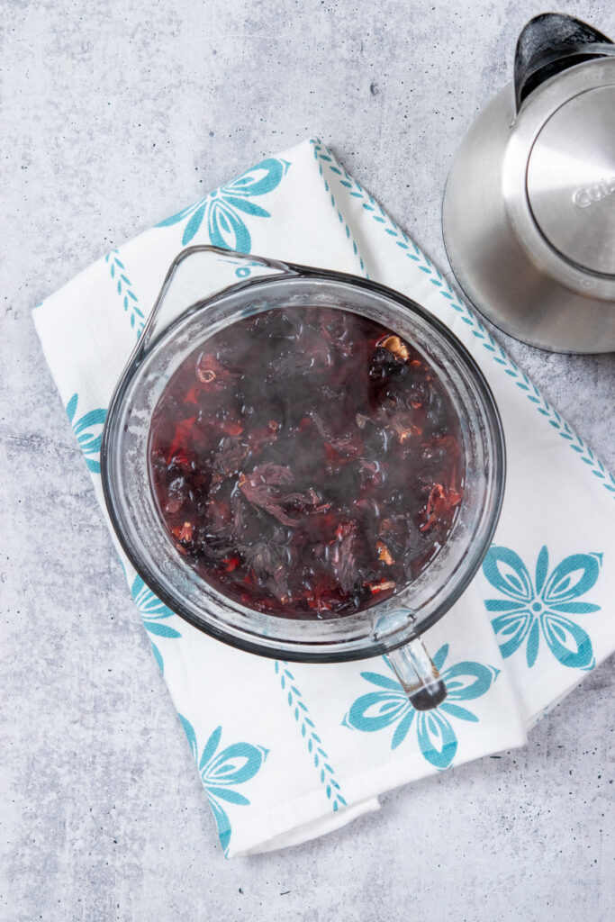 A large 4-cup heat-safe glass measuring cup with spout filled with hibiscus leaves steeping in hot water. Next to the tea is an electric kettle which was used to heat the water.