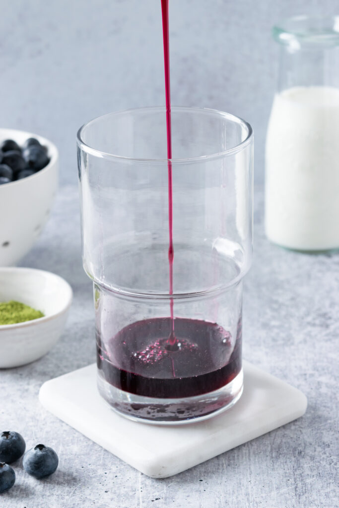 Stream of blueberry syrup being poured into a clear drinking glass, with matcha powder and blueberries in the background.