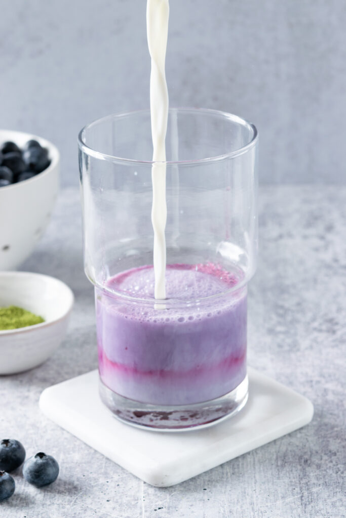 Milk being poured into blueberry syrup in a clear glass, with bowls of matcha and blueberries behind.