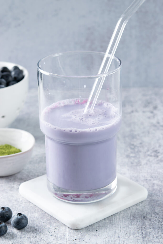 Blueberry syrup and milk stirred together with a glass straw in a clear glass to create a pale purple base.