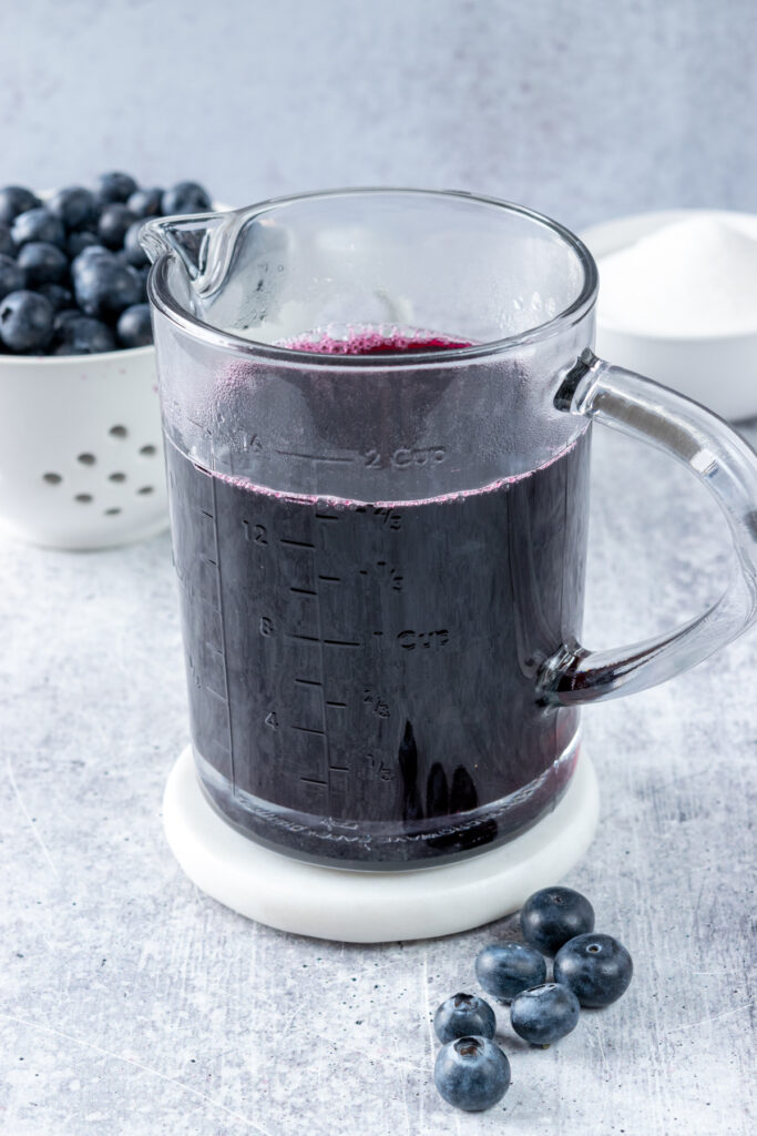 Two cups of homemade blueberry syrup for drinks in a glass measuring cup. The syrup is in front of a berry bowl filled with fresh blueberries, and a small bowl of sugar.