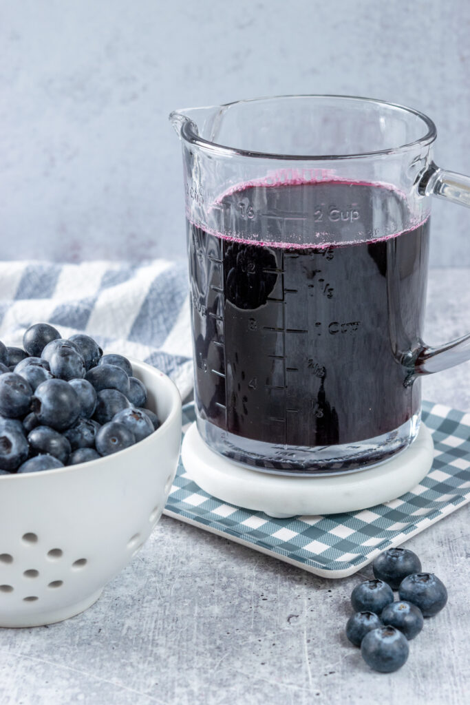 Fresh blueberries in a berry bowl which is next to a glass measuring cup of homemade blueberry syrup. In the foreground are a few loose blueberries on the table and the simple syrup is on a white coaster which is on a blue checked plate.