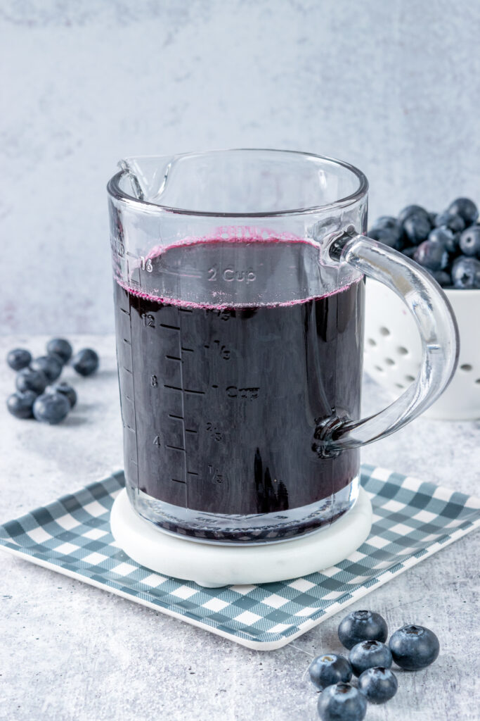 A jar of homemade blueberry simple syrup for drinks with a berry bowl in the background and a few loose blueberries strewn about on the table.