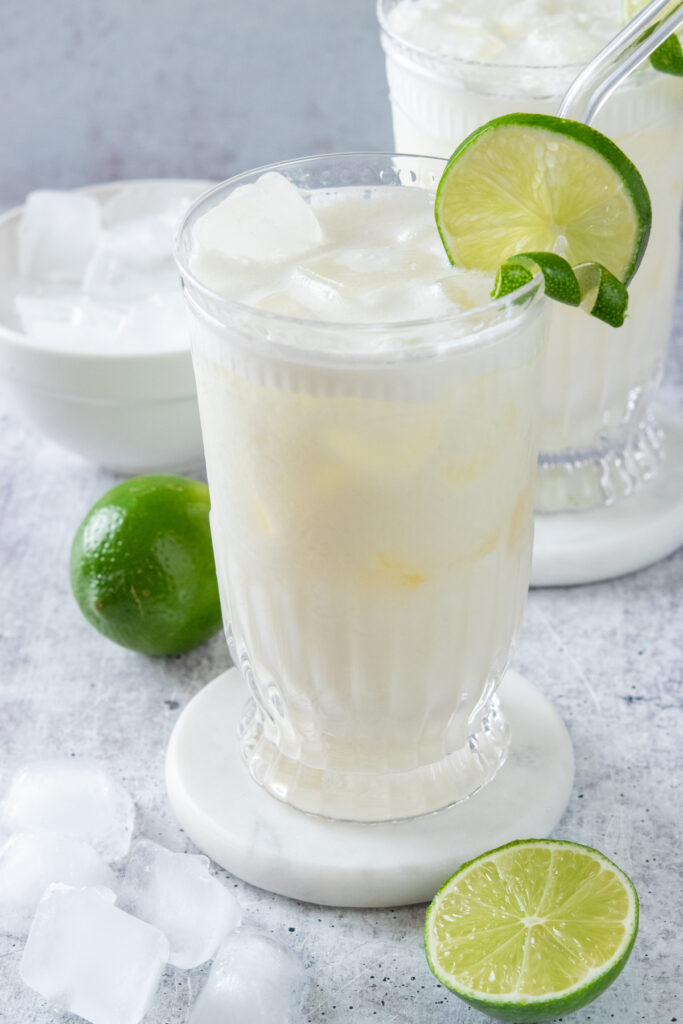 Two tall glasses of ice cold Brazilian lemonade with lime twists and straws. In the foreground are some ice cubes and half a lime. In the background is a full lime and a bowl of ice cubes.