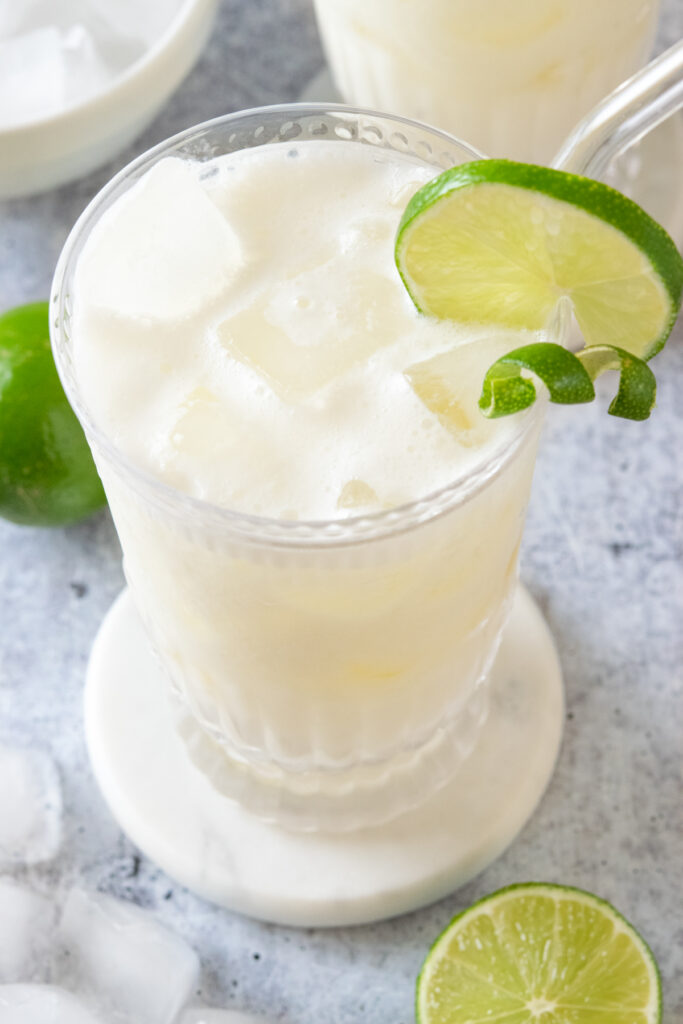 A top-down view of a creamy refreshing limeade drink that's garnished with a round slice of lime and a lime twist. This Brazilian lemonade is surrounded on the table with ice cubes and limes.