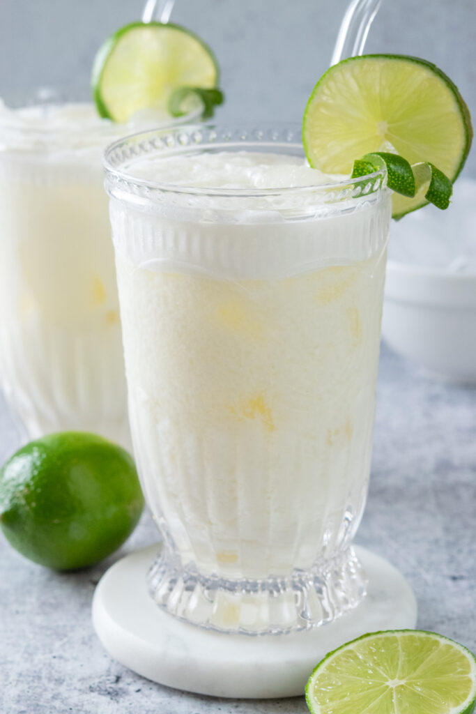 Two glasses of Brazilian lemonade over ice. Both drinks are garnished with limes and there is a full lime and a bowl of ice cubes in the background.