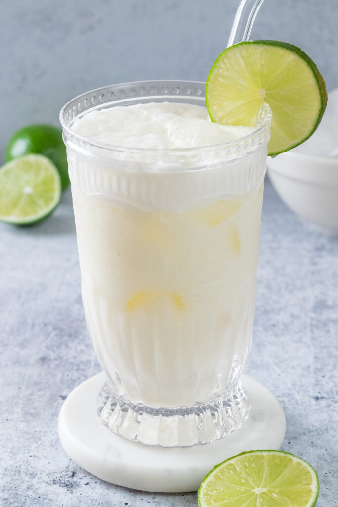 Brazilian lemonade over ice in a glass that's garnish with a slice of lime and has a straw in it.