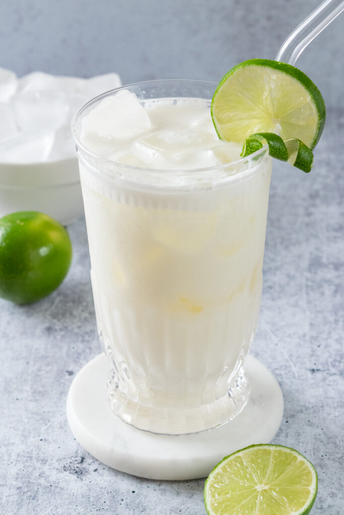 A tall glass of Brazilian limeade garnished with a lime twist and round slice of lime. There is a lime and bowl of ice cubes in the background.