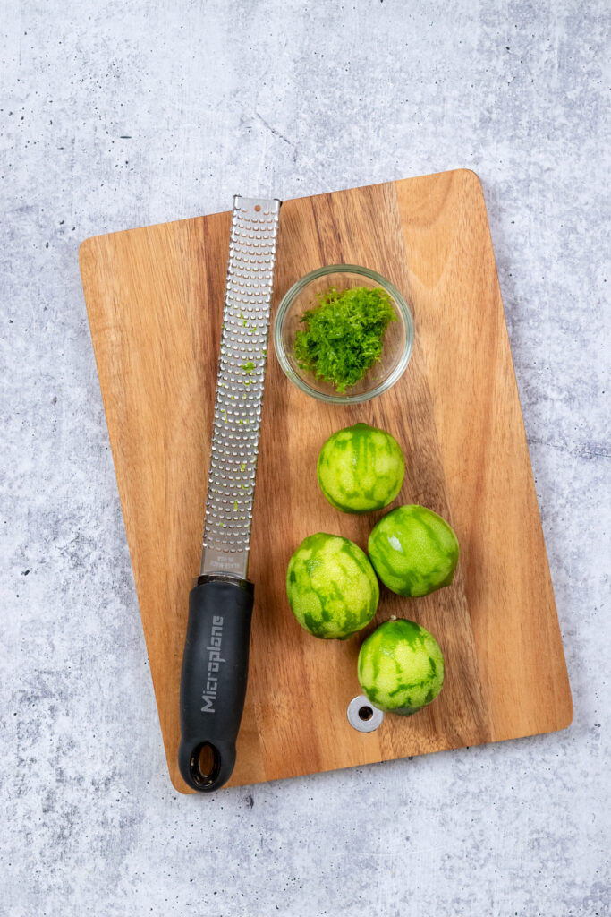 An overhead view of four limes on a cutting board that have been zested. The lime zest is in a small bowl and the zester is pictured, as well.