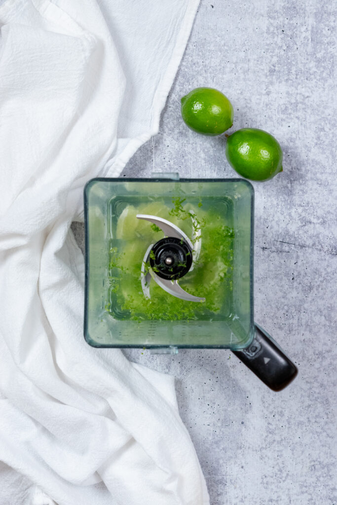 Overhead view of a blender with peeled limes, lime zest and water before being blended.