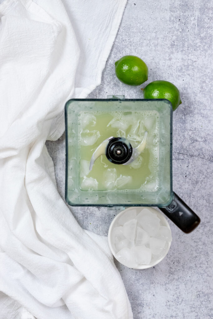An overhead view of a blender that has the lime water returned to it, and condensed milk and a handful of ice cubes add in, ready to blend.