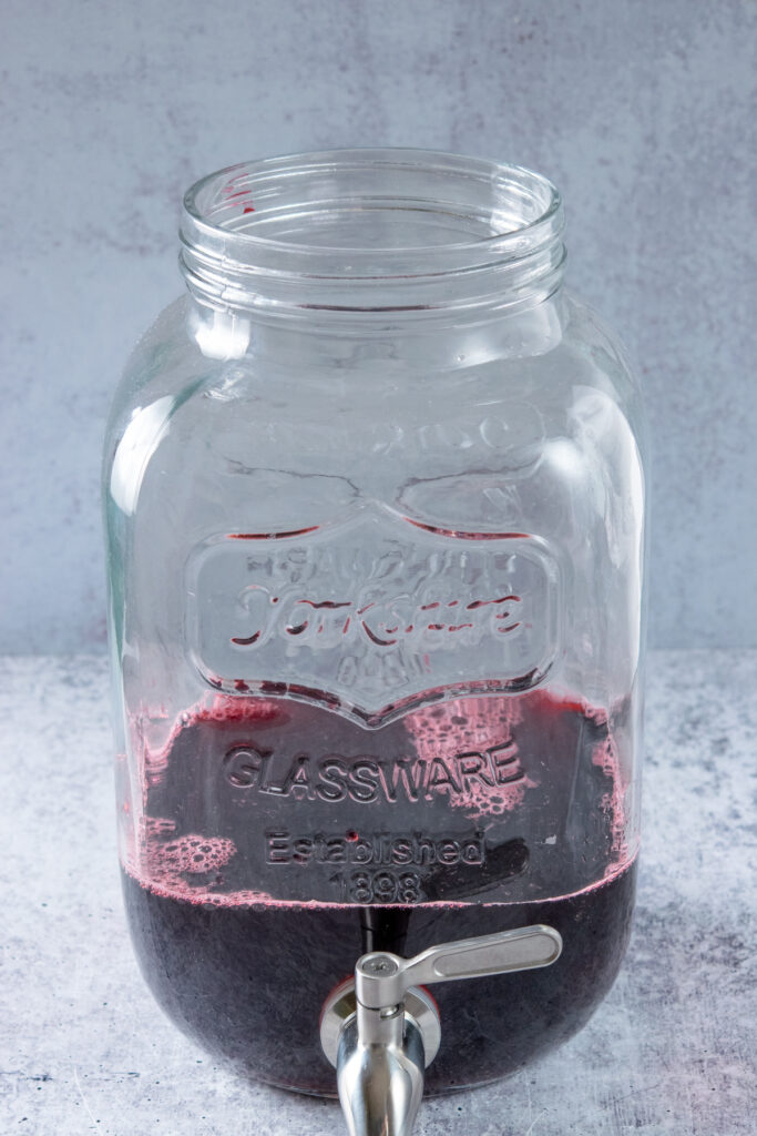 Making a big-batch of homemade hibiscus iced tea by first adding concentrated hibiscus tea to a glass gallon container which has a spigot for easy serving.