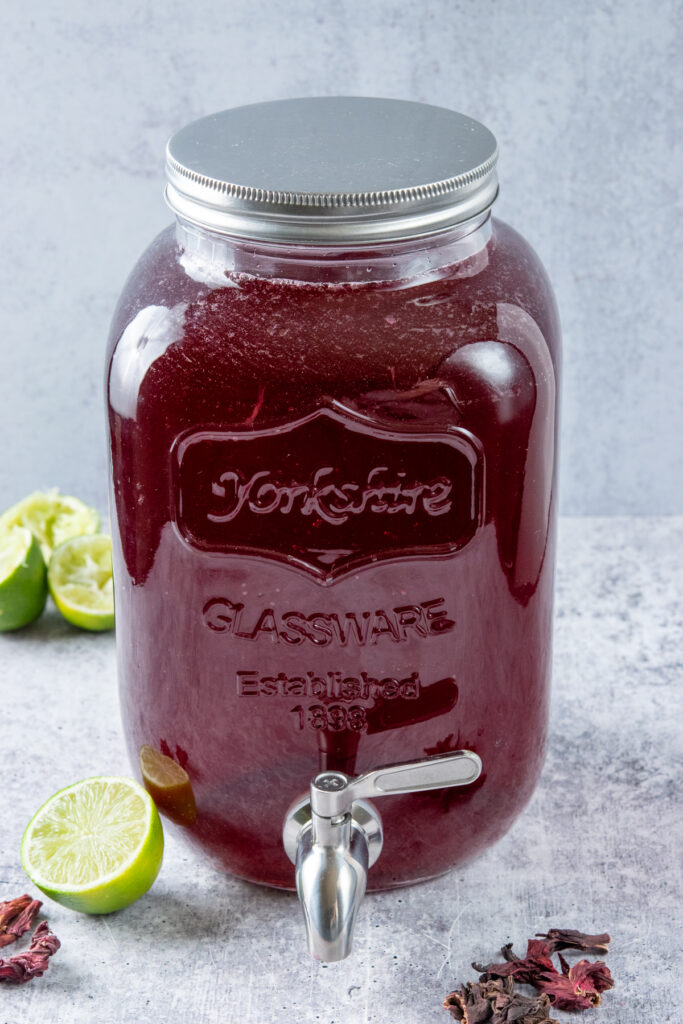 A large glass gallon container that has a spigot on it for easy serving, that's filled with the most refreshing homemade hibiscus iced tea. The glass jar is surrounded by limes and dried hibiscus flowers.