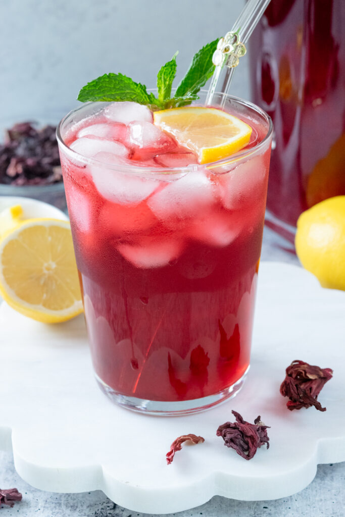A refreshing glass of iced hibiscus lemonade that's topped with a sprig of mint and lemon slice. The drink is next to a pitcher full of the ruby red lemonade, as well as a full lemon and small bowl of dried hibiscus flowers.