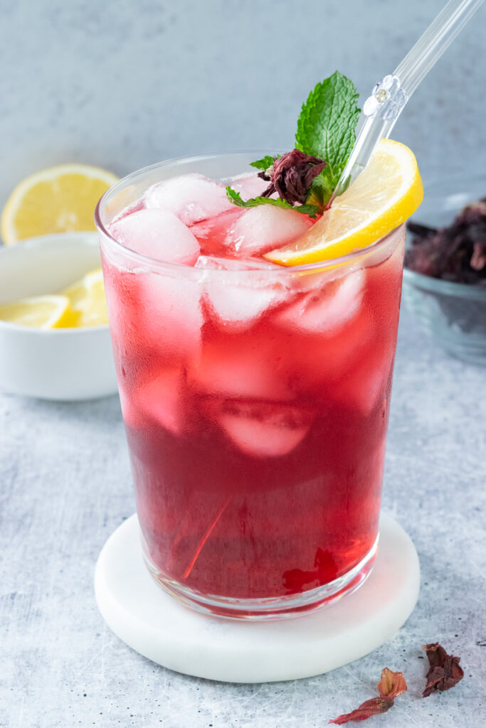 A glass of homemade hibiscus lemonade that's filled with ice and garnished with a slice of lemon, mint leaves and a small piece of dried hibiscus flower. The drink has a glass straw in it that is decorated with a glass flower that resembles hibiscus. Behind the lemonade drink is a small bowl filled with lemon slices and a small glass bowl filled with dried hibiscus flowers.