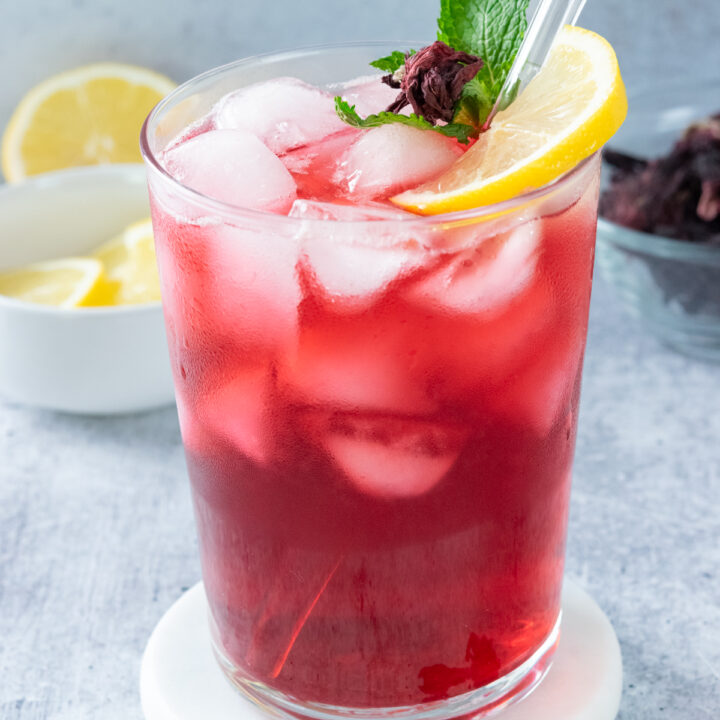 A glass of homemade hibiscus lemonade that's filled with ice and garnished with a slice of lemon, mint leaves and a small piece of dried hibiscus flower. The drink has a glass straw in it that is decorated with a glass flower that resembles hibiscus. Behind the lemonade drink is a small bowl filled with lemon slices and a small glass bowl filled with dried hibiscus flowers.