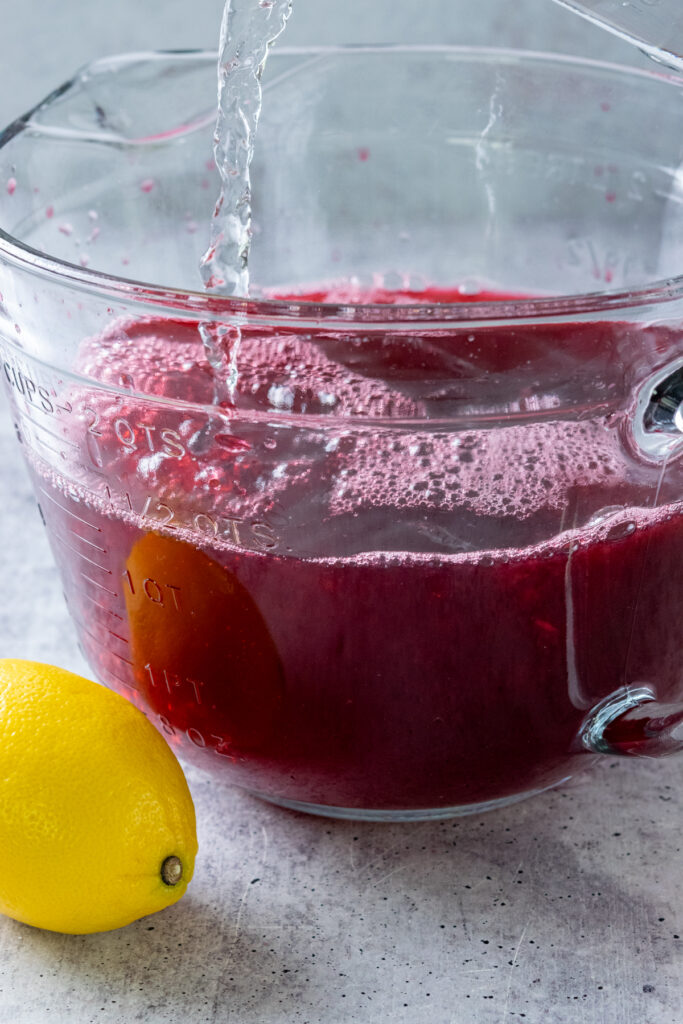 Pouring cold water into concentrated hibiscus tea to make homemade hibiscus lemonade.