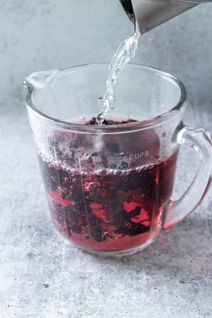 Making concentrated hibiscus tea by pouring hot water over dried hibiscus flowers in a heatproof glass measuring cup.