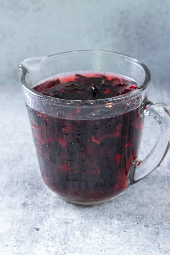 Hibiscus steeping in hot water. The water has turned a deep ruby red and you can see the dried flowers floating in it.