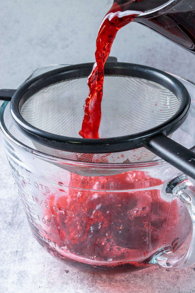 Pouring the concentrated hibiscus tea, through a fine-mesh strainer into a large glass pitcher.