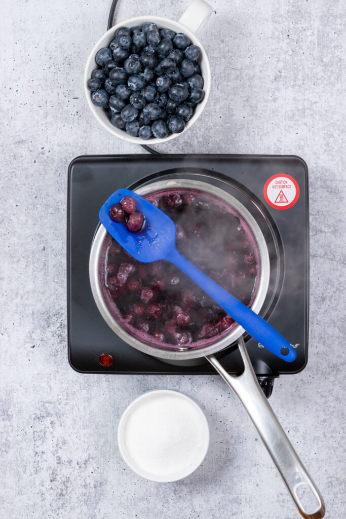 A spoon resting on a saucepan of cooking blueberry syrup. The spoon has three cooked blueberries in it that are almost pink.