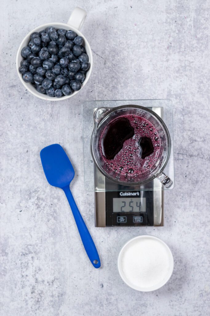 A glass measuring cup filled with the concentrated blueberry juice on  a scale showing that it weighs 254 grams, which is then the amount of sugar to use.