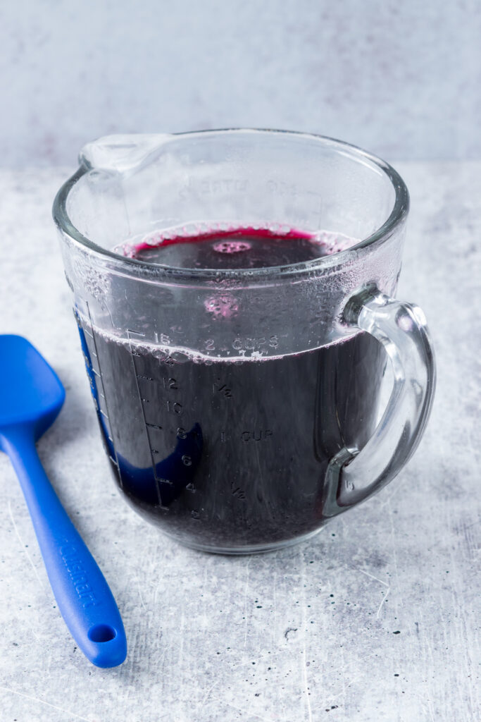 Two cups of hot blueberry syrup in a glass measuring cup ready to be cooled down.