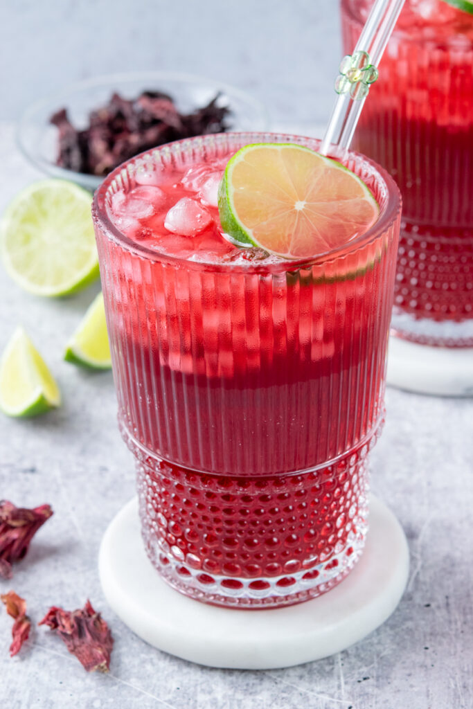 Two ribbed glasses filled with red hibiscus iced tea and lots of ice. Each drink is garnished with lime and has a glass straw with a glass flower on it inserted into the iced tea. In the background is half a lime and a small bowl filled with dried hibiscus flowers. The drinks look super refreshing and ice cold.