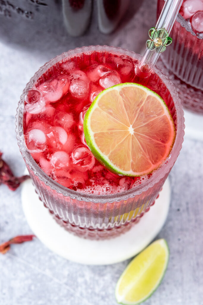 An overhead photo of a refreshing glass of homemade hibiscus iced tea. The drink is filled with mini sphere ice cubes, is garnished with a round lime slice and has a glass straw that's embellished with a glass flower inserted into it. There are a few dried hibiscus petals scattered around the table that the drink is sitting on, as well as a lime wedge.
