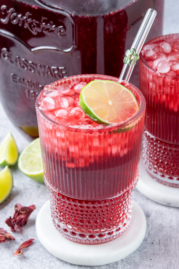 Two glasses filled to the brim with homemade hibiscus iced tea. The drinks are in front of a gallon container of the hibiscus tea. Each drink is garnished with lime and there are a few lime wedges and dried hibiscus petals surrounding the drinks.