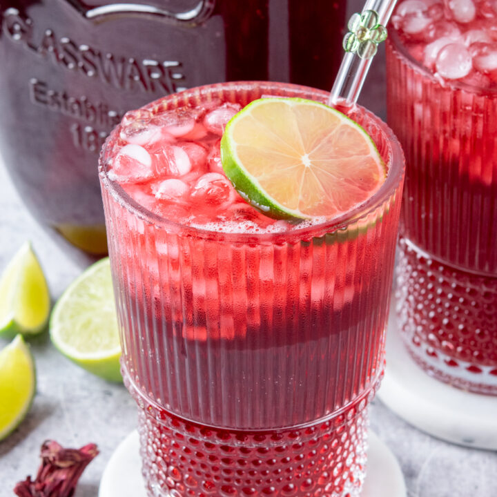 Two glasses filled to the brim with homemade hibiscus iced tea. The drinks are in front of a gallon container of the hibiscus tea. Each drink is garnished with lime and there are a few lime wedges and dried hibiscus petals surrounding the drinks.