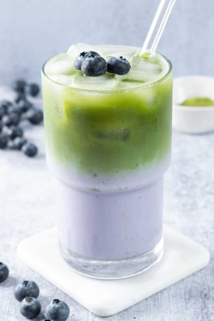 An iced blueberry matcha latte in a tall glass with a glass straw in it and three blueberries on top. The drink is surrounded by loose blueberries on the table and a small bowl of matcha powder.