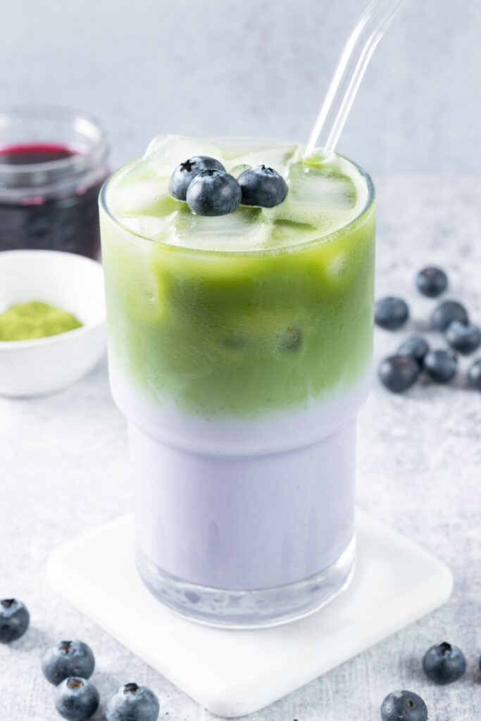 An iced blueberry matcha latte in a tall glass that shows the layers in the drink. The latte has a glass straw in it and three blueberries on top. In the background is a jar of blueberry syrup, a bowl of matcha powder and some blueberries.