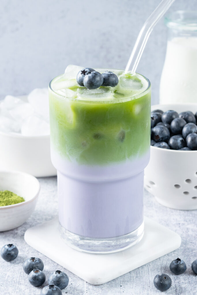A layered iced blueberry matcha latte with three blueberries on top. The drink is in front of a bowl of matcha powder, some ice cubes, blueberries in a berry bowl, and a pitcher of milk.