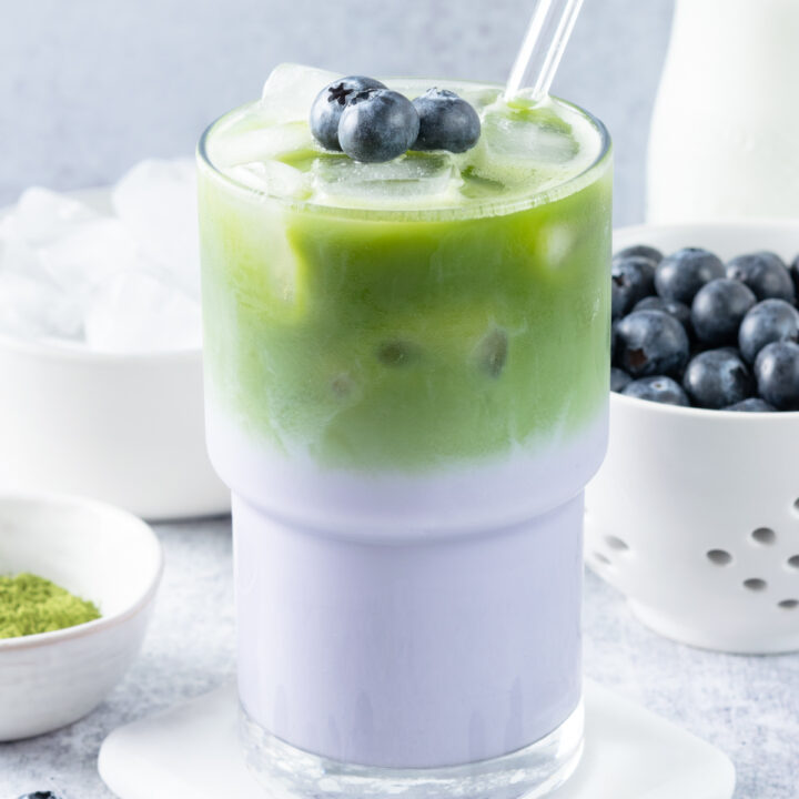 A layered iced blueberry matcha latte with three blueberries on top. The drink is in front of a bowl of matcha powder, some ice cubes, blueberries in a berry bowl, and a pitcher of milk.