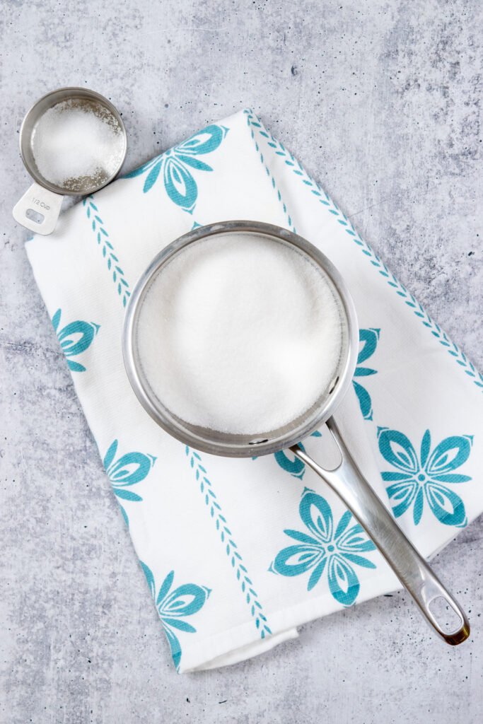 A small saucepan with a cup of sugar in it to make simple syrup. The saucepan is on a white towel with teal-colored flowers on it.