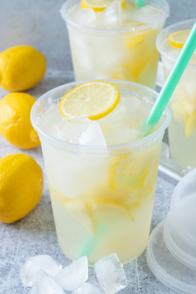 Three large deli cups filled with homemade state fair lemonade. There are three whole lemons and three lids next to the drinks. Each lemonade has lots of iced and lemons in it and a slice of lemon on top.