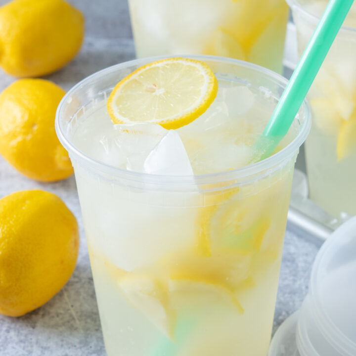 Three large deli cups filled with homemade state fair lemonade. There are three whole lemons and three lids next to the drinks. Each lemonade has lots of iced and lemons in it and a slice of lemon on top.
