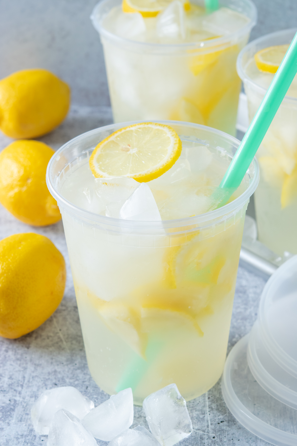 Three large deli cups filled with homemade state fair lemonade. There are three whole lemons and three lids next to the drinks. Each lemonade has lots of iced and lemons in it and a slice of lemon on top.