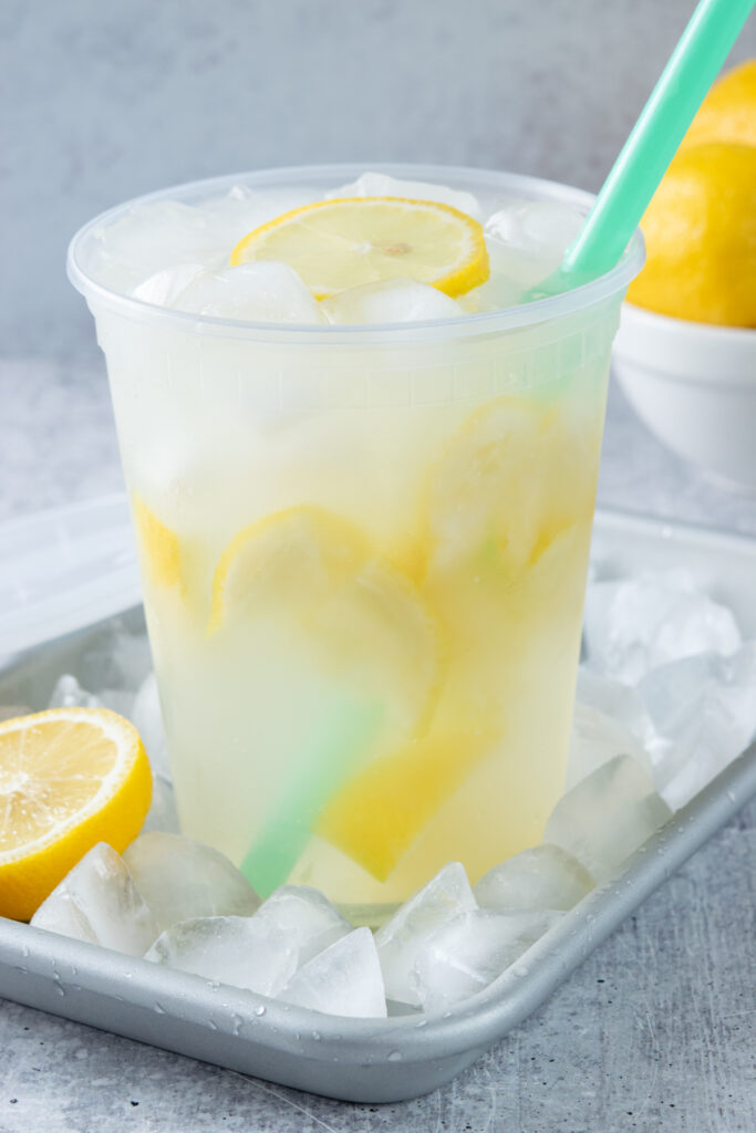 A refreshing cup of homemade smashed lemonade made in a deli container for serving. The drink is filled with pieces of lemon and lots of ice. There is a slice of lemon floating on top.